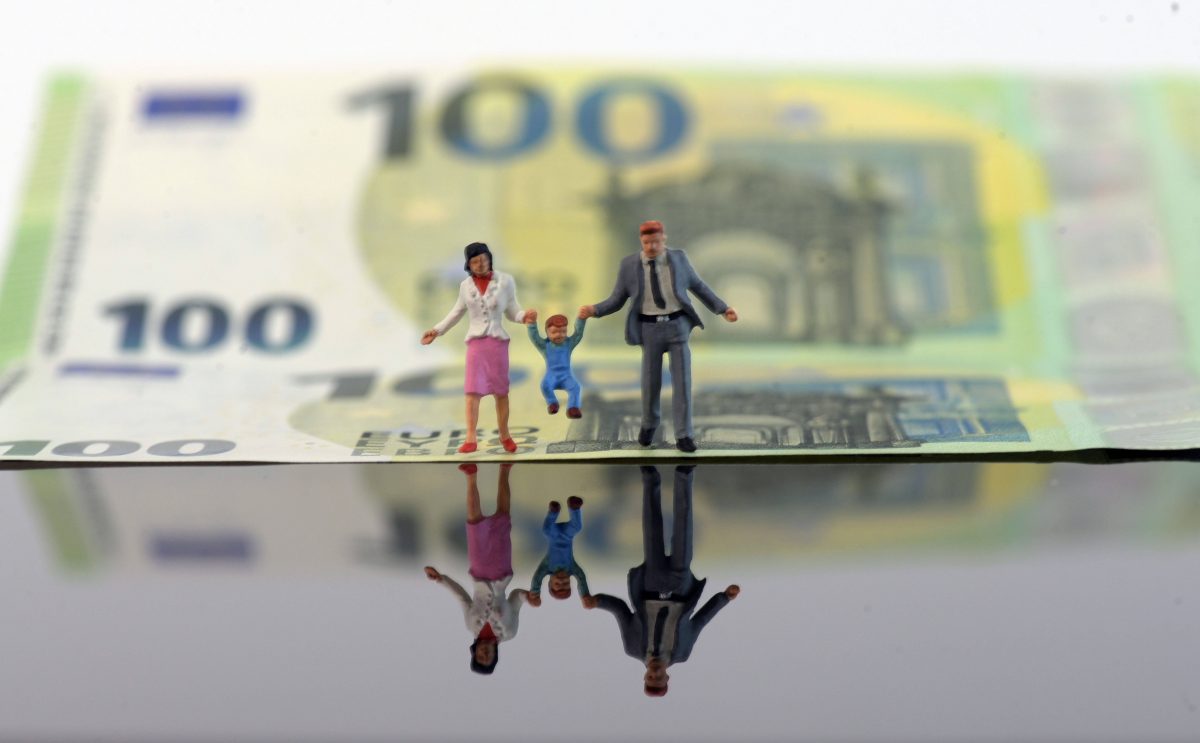 Figuren von zwei Erwachsenen halten ein Kind an ihren Händen. Im Hintergrund ein 100-Euro-Schein.