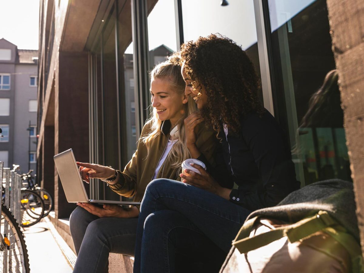 Zwei junge Frauen sitzen vor einer Universitätsbibliothek vor dem Laptop