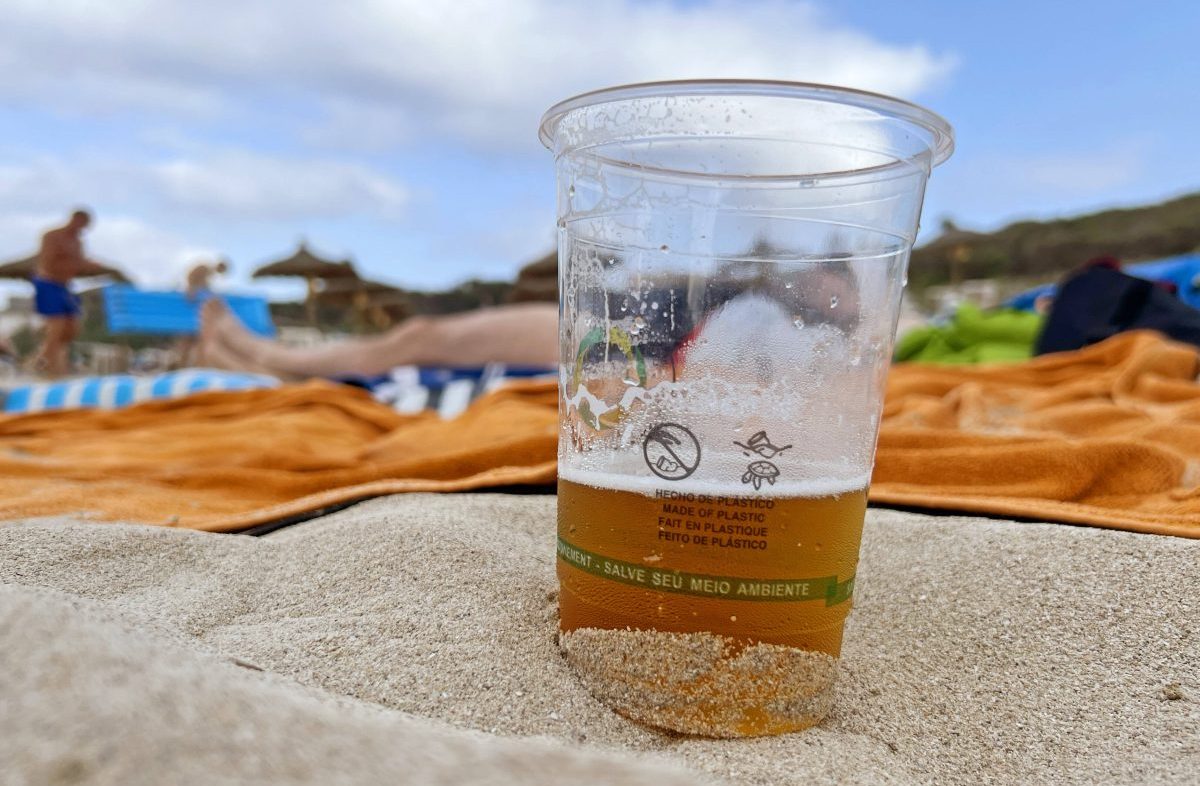 halbgefüllter Bierbecher am Strand von Mallorca, Urlauber auf Handtüchern im Hintergrund