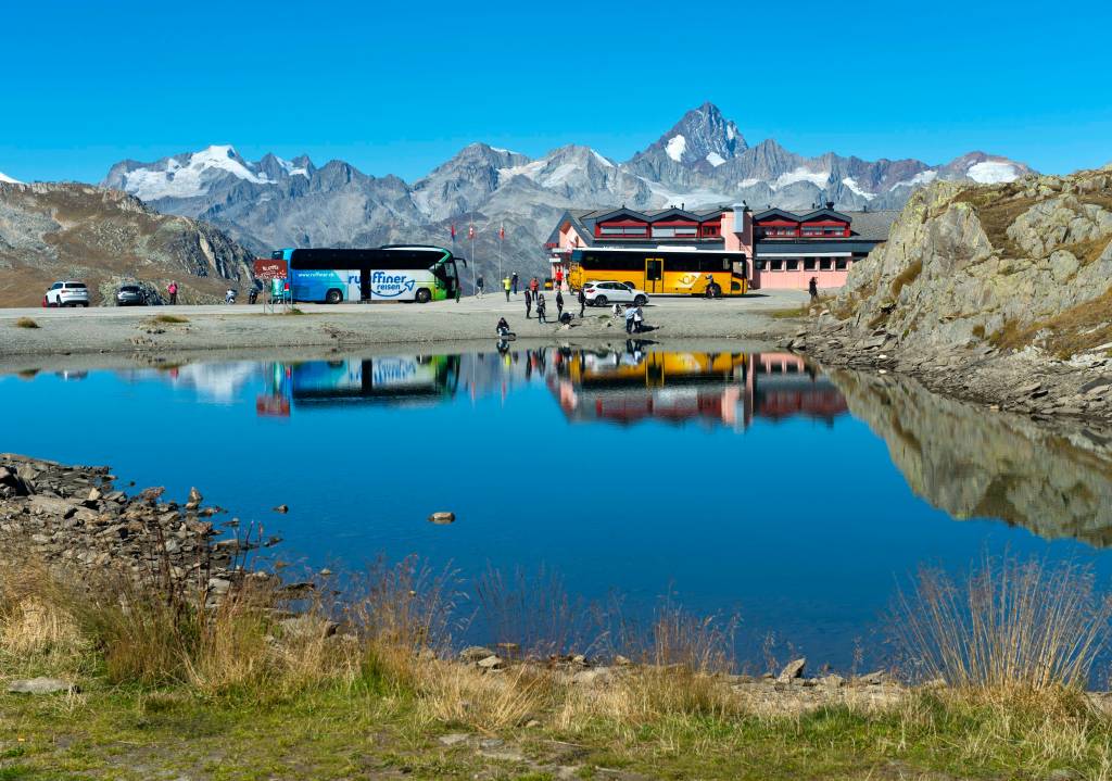 Reisebus mit Touristen und gelber Linienbus der Schweizer PostAuto AG auf dem Nufenenpass, hinten die Bergkette der Walliser Alpen.