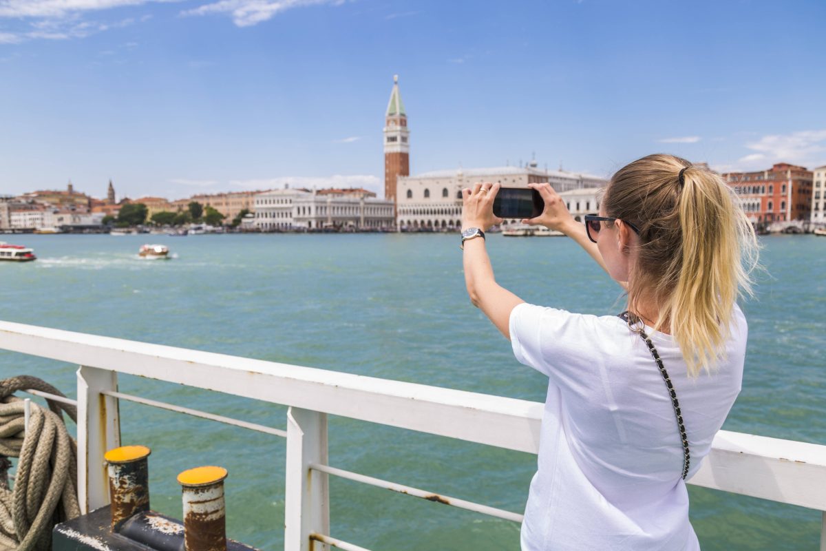 Eine Frau ist auf einem Schiff und steht an der Reling. Sie macht mit ihrem Handy ein Foto von einer Sehenswürdigkeit.