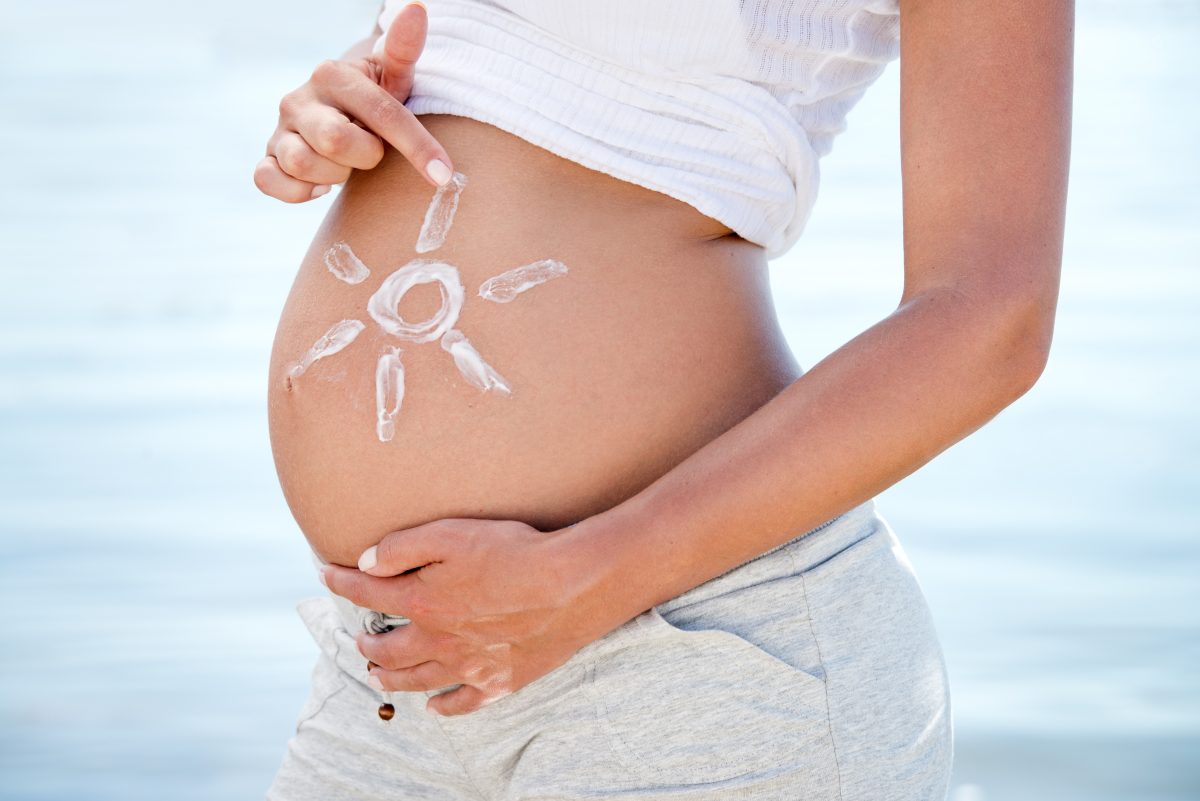 Schwangerschaftsbauch von der Seite. Die Person hält ihre Hände an den Bauch. Mit einer Hand malt sie eine Sonne mit Sonnencreme auf die Seite vom Bauch.