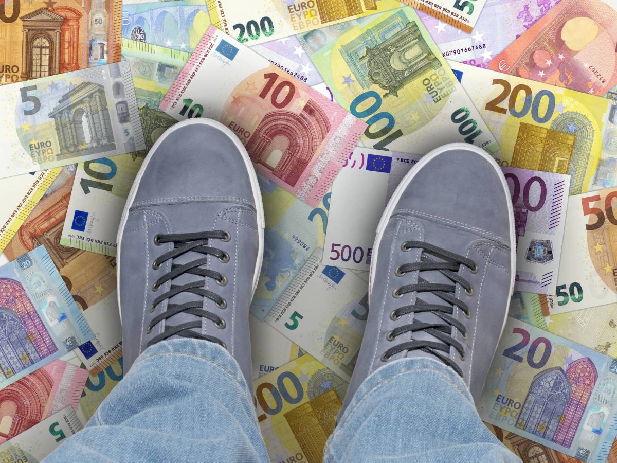 Ein Mensch steht mit beiden Füßen auf Euro-Geldscheinen, die auf dem Boden verteilt liegen.