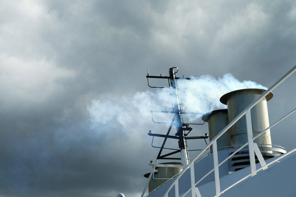 Der Auspuff eines Kreuzfahrtschiffes, aus dem gerade Abgase in Form von Rauchwolken entweichen. Im Hintergrund ist ein grauer Himmel zu sehen.