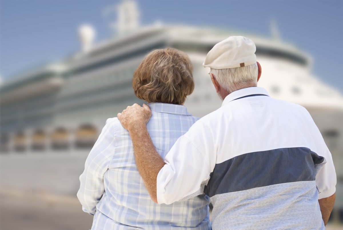 Ein Senioren-Pärchen steht Am in Arm vor einem Kreuzfahrtschiff. Der Mann hat eine Mütze auf uns den linken Arm auf der Schulter seiner Frau.