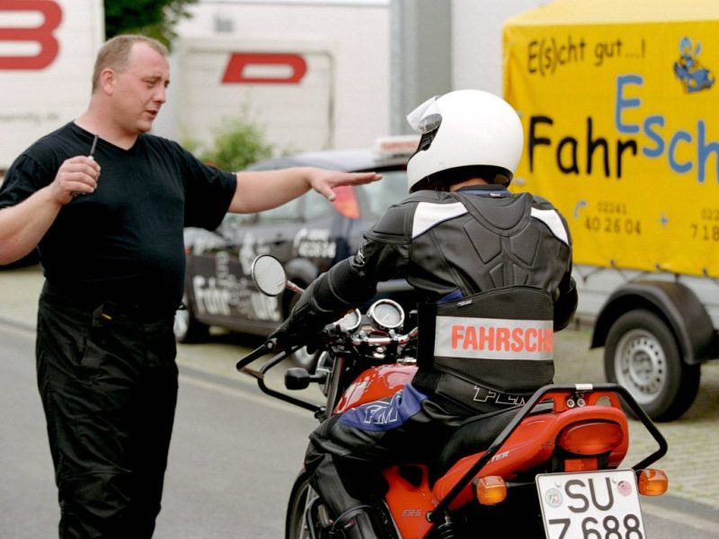 Im Rücken zum Bild sitzt in Fahrschüler auf einem roten Motorrad. Er hat einen weißen Helm auf und eine Jacke auf der in rot "Fahrschule" steht. Vor im zeigt ein Mann mit einem Finger auf ihn. Er trägt eine schwarze Hose und ein schwarzes T-Shirt.