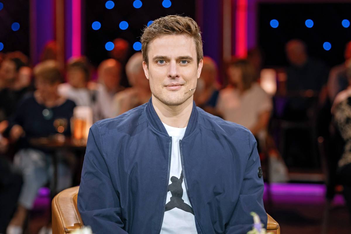 Auf dem Bild ist Tagesschau-Moderator und Schriftsteller Constantin Schreiber. Er sitzt in einem Fernsehstudio. Er trÃ¤gt ein weiÃŸes T-Shirt und darÃ¼ber eine blaue Bomberjacke. Er hat braune hochgegelte Haare und an seiner Wange ein Mikrofon angebracht.