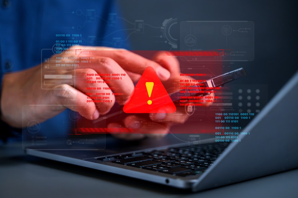 Eine Hand hält über einem Notebook ein Smartphone. Ein Warnsignal leuchtet auf.