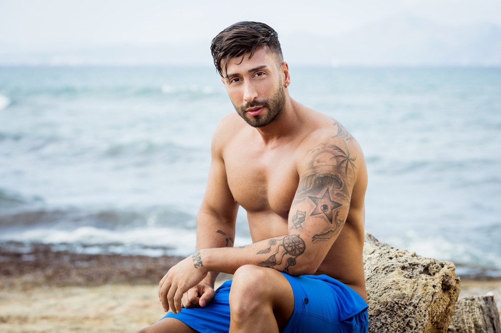 Bobby Chambers sitzt in blauer Badehose auf einem Stein am Meer